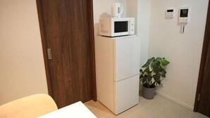 A compact room with a white refrigerator, a microwave on top, a potted plant, and a door with a wooden frame. - Ashe Maison Tennoji West II (Osaka)