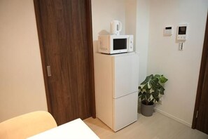 Ein kleines Zimmer mit einem weißen Kühlschrank, einer Mikrowelle darauf, einer Blumentopf-Pflanze und einer Tür mit Holzrahmen.