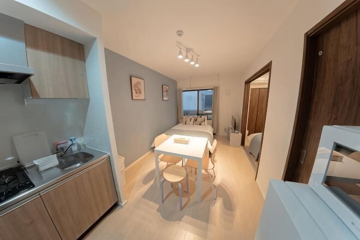A compact kitchen with a stove, sink, and white table with chairs. A window provides natural light to the adjacent bedroom.