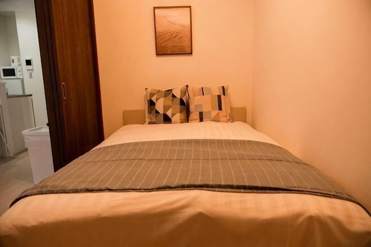 A bed with a patterned bedspread, a wall-mounted artwork, and a door leading to a kitchenette.