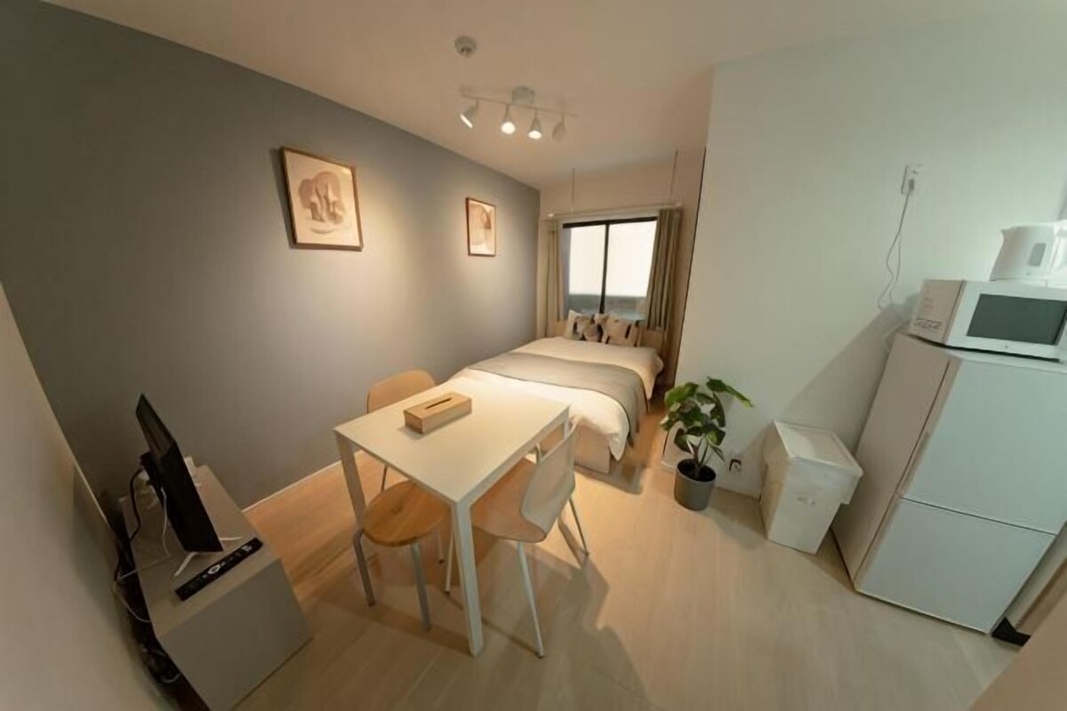 A compact hotel room with a bed, a desk, a chair, a small kitchenette, and a potted plant.