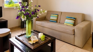 Een woonkamer met een beige bank, een glazen vaas met bloemen, een kaars en een dienblad met boeken en een afstandsbediening.