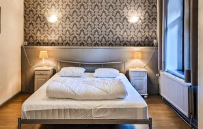 Ein ordentlich bezogenes Bett in einem Zimmer mit ornamentalem Wandpapier, zwei Nachttischlampen, einem Fenster und einer Heizung.