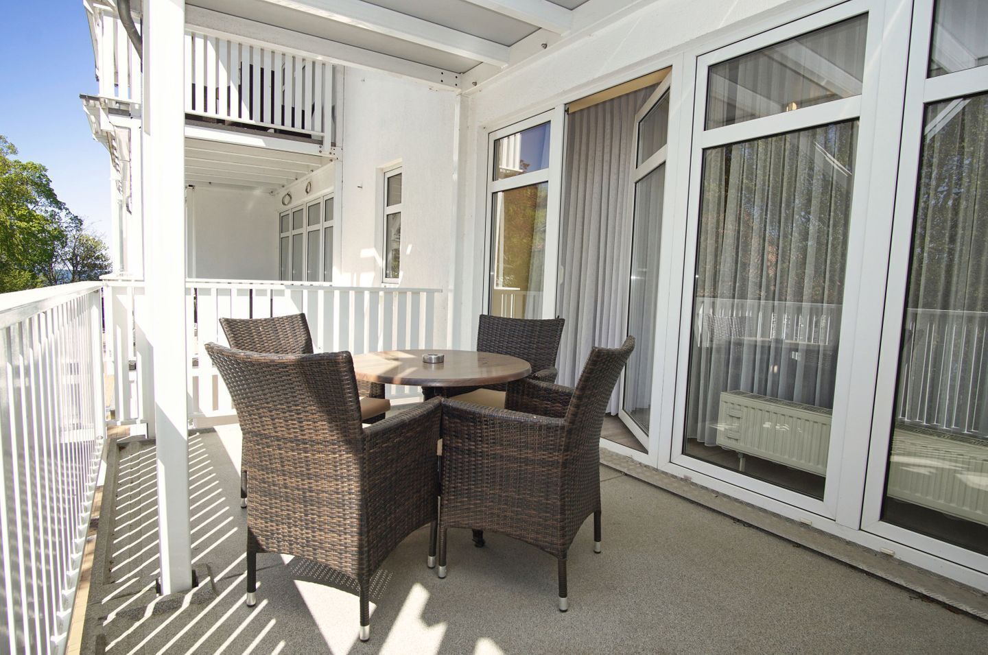 Ein Balkon mit Korbstühlen, einem Tisch, großen Fenstern und einem weißen Geländer.