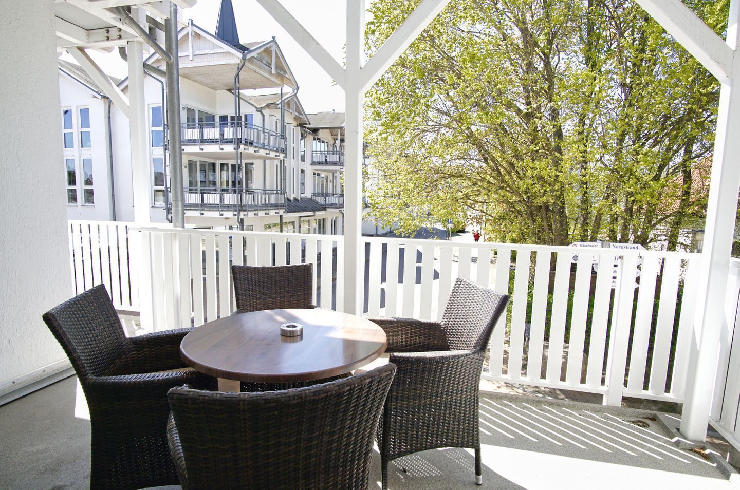 A covered outdoor seating area with wicker chairs and a round table, white railings, and a view of buildings and trees.
