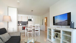 Ett modernt vardagsrum med en soffa, ett soffbord i glas, en tv och ett kök i bakgrunden.