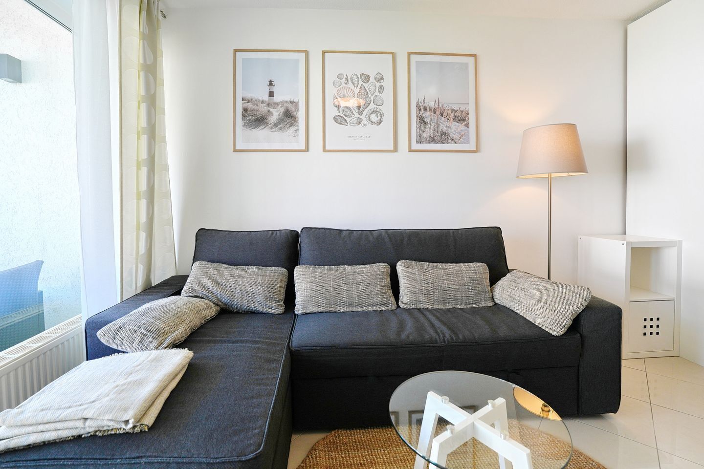 A modern living room with a dark grey sofa, a glass coffee table, and a lamp. There are three framed pictures on the wall.