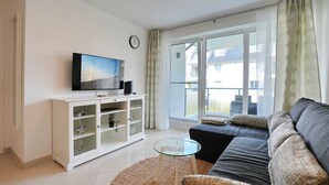 Ett vardagsrum med en soffa, ett soffbord i glas, en tv som står på ett skåp och en skjutdörr i glas som leder ut till uteplatsen.