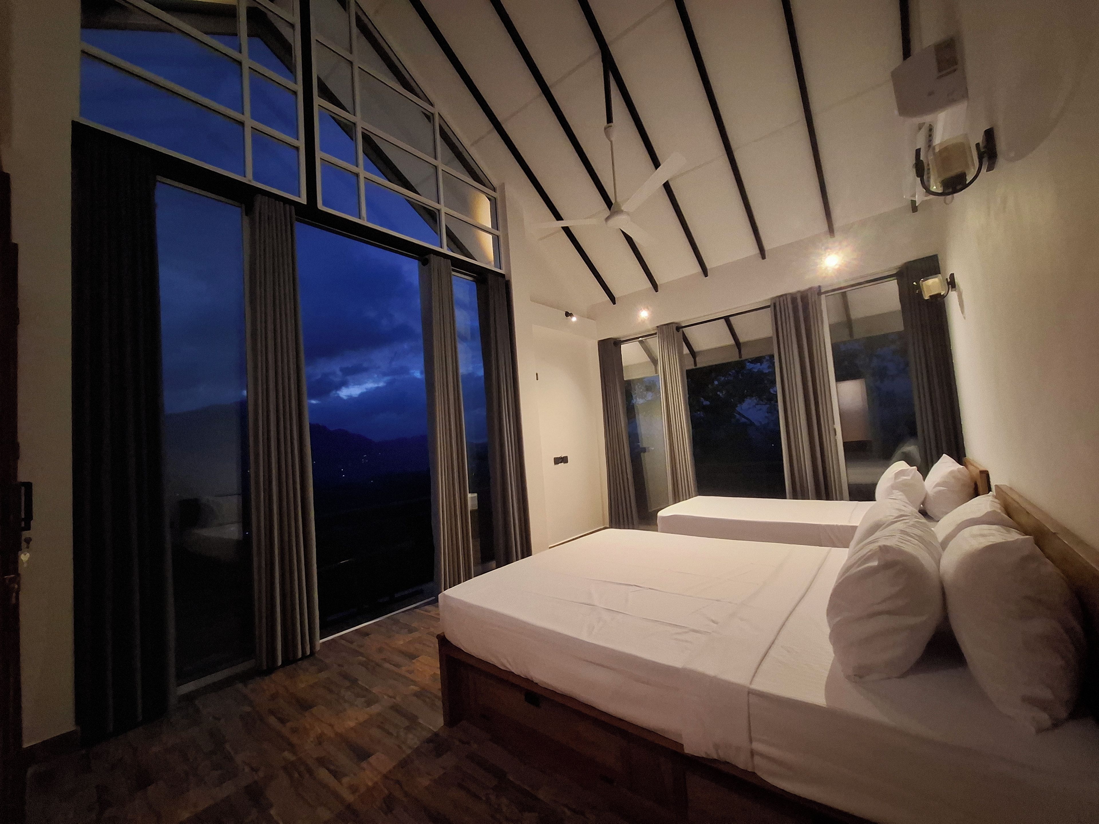 Un dormitorio moderno con una cama grande, un ventilador de techo y vistas del cielo nocturno a través de amplios ventanales.