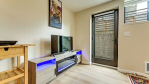 A modern living room with a flat-screen TV, wooden entertainment unit, and a sliding glass door leading outside. - Walk to Seward Park! Pet-Friendly Apt w/ Turf Yard (Seattle)