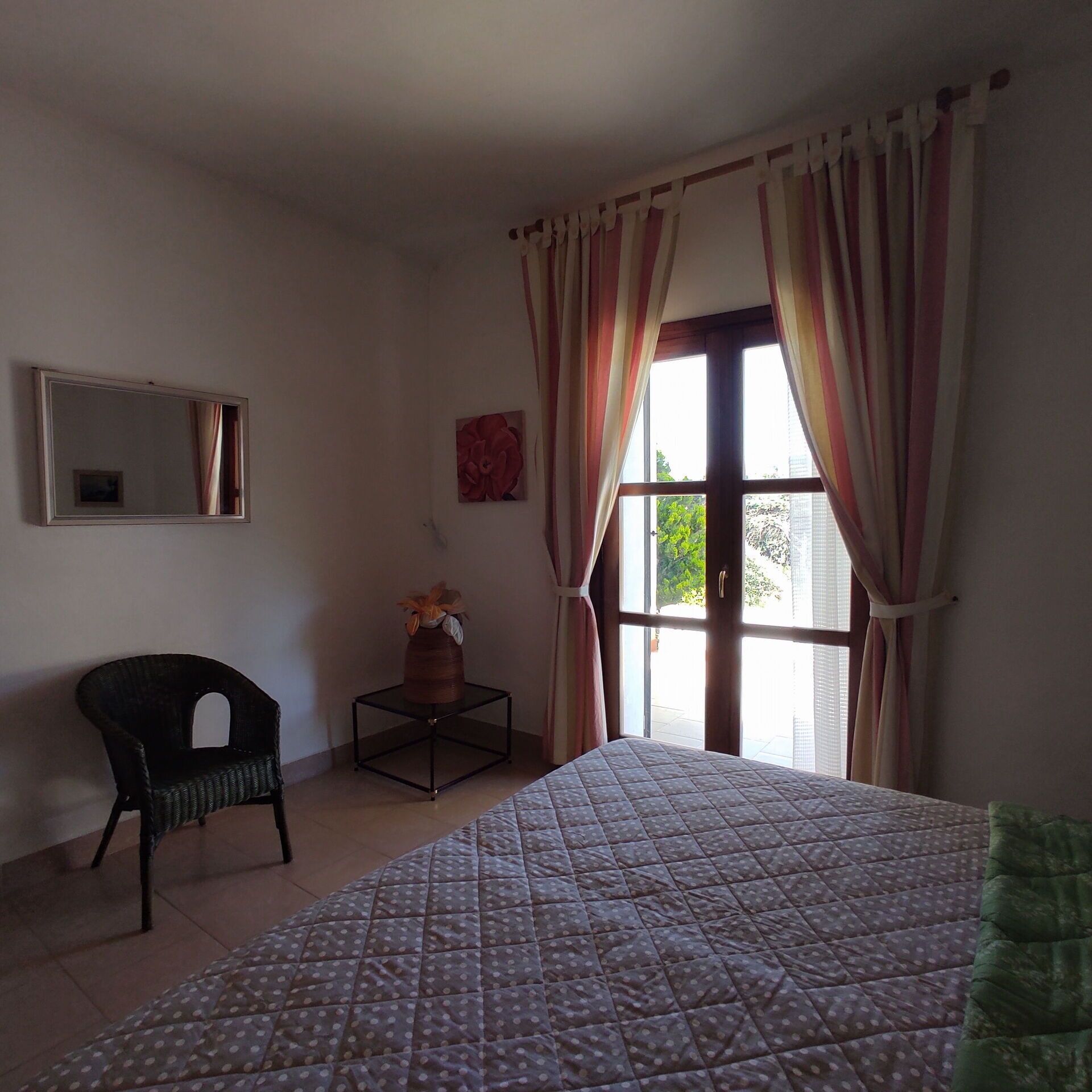 Una camera da letto con un letto, una sedia, un tavolino con un vaso, uno specchio e una finestra con tende.