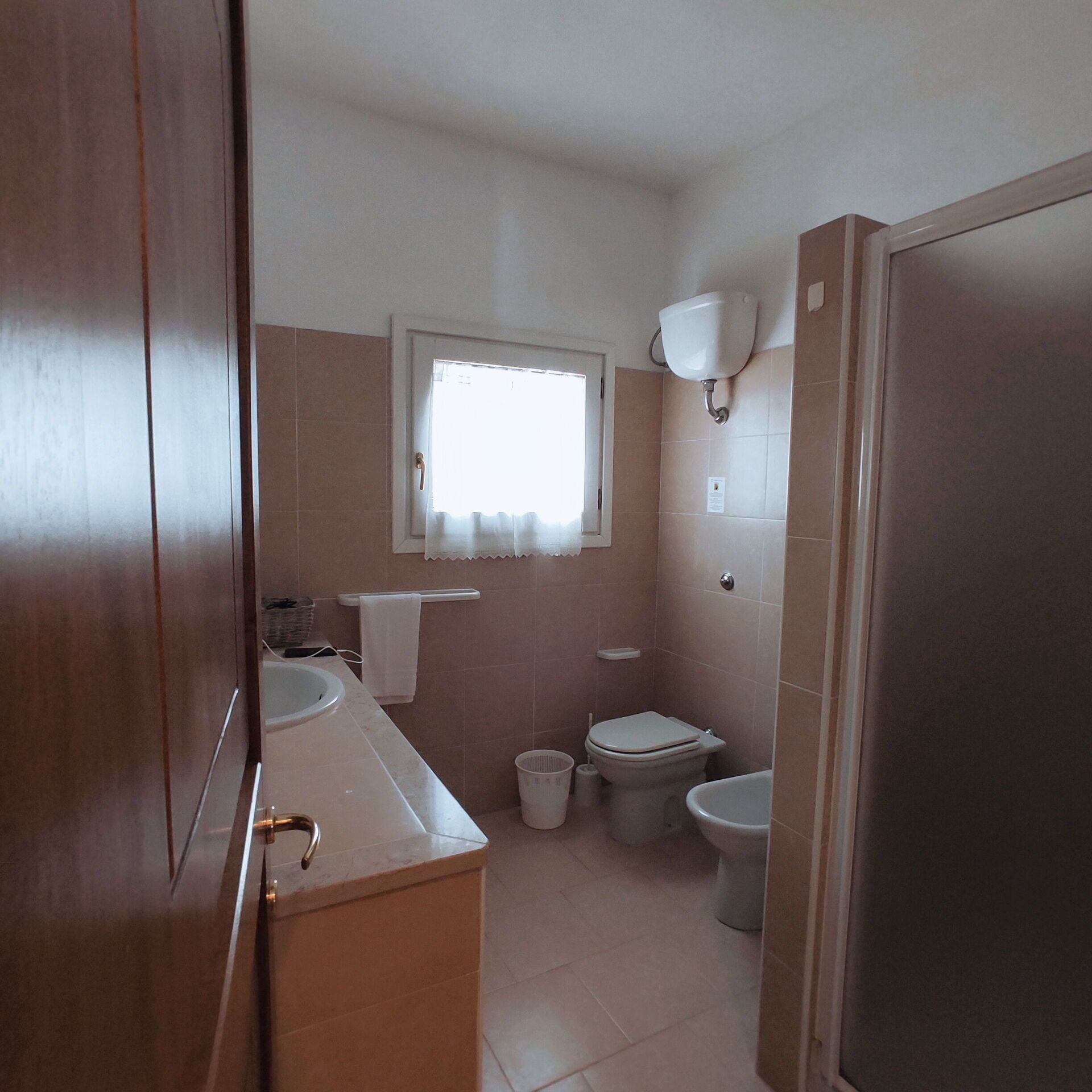 A bathroom with a toilet, bidet, sink, and a window with a curtain.