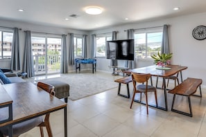 Una sala moderna con sofá, una mesa de comedor con bancos, televisor de pantalla plana, mesa de futbolín y un balcón con cortinas.