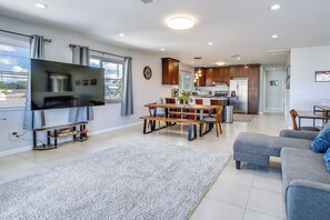 A modern living room with a flat-screen TV, a dining area with a wooden table and chairs, a grey sofa, and a kitchen with dark wooden cabinets and stainless steel appliances. - Walk to Sand! Ewa Beach Group Retreat (Ewa Beach)