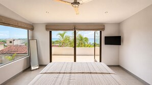 Un dormitorio con una cama grande, ventilador de techo, televisor en la pared y un balcón con vista al mar.