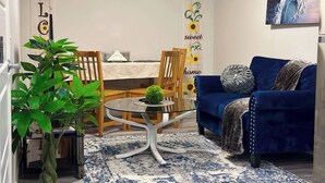 A living room with a blue sofa, a glass coffee table, a dining area with a table and chairs, and a wall with a 'WELCOME' sign and a sunflower banner. - Cozy Private Suite Near Stoney & Deerfoot Trails (Calgary)