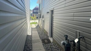 A narrow pathway between two buildings with a metal door and outdoor water pipes. - Cozy Private Suite Near Stoney & Deerfoot Trails (Calgary)