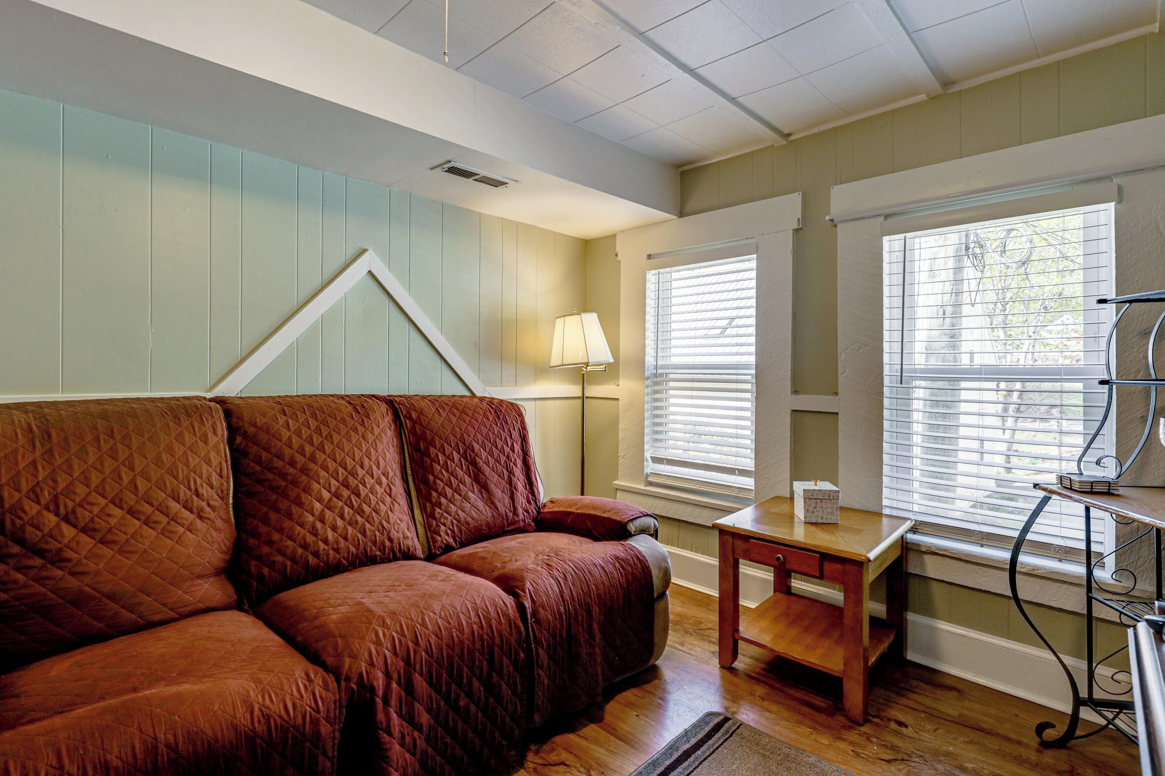 A room with a brown quilted sofa, a small wooden table, a lamp, and a window with blinds.