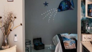 Una habitación con un sofá azul, una cama con una manta estampada, un mural de peces en la pared y un ventilador de techo.