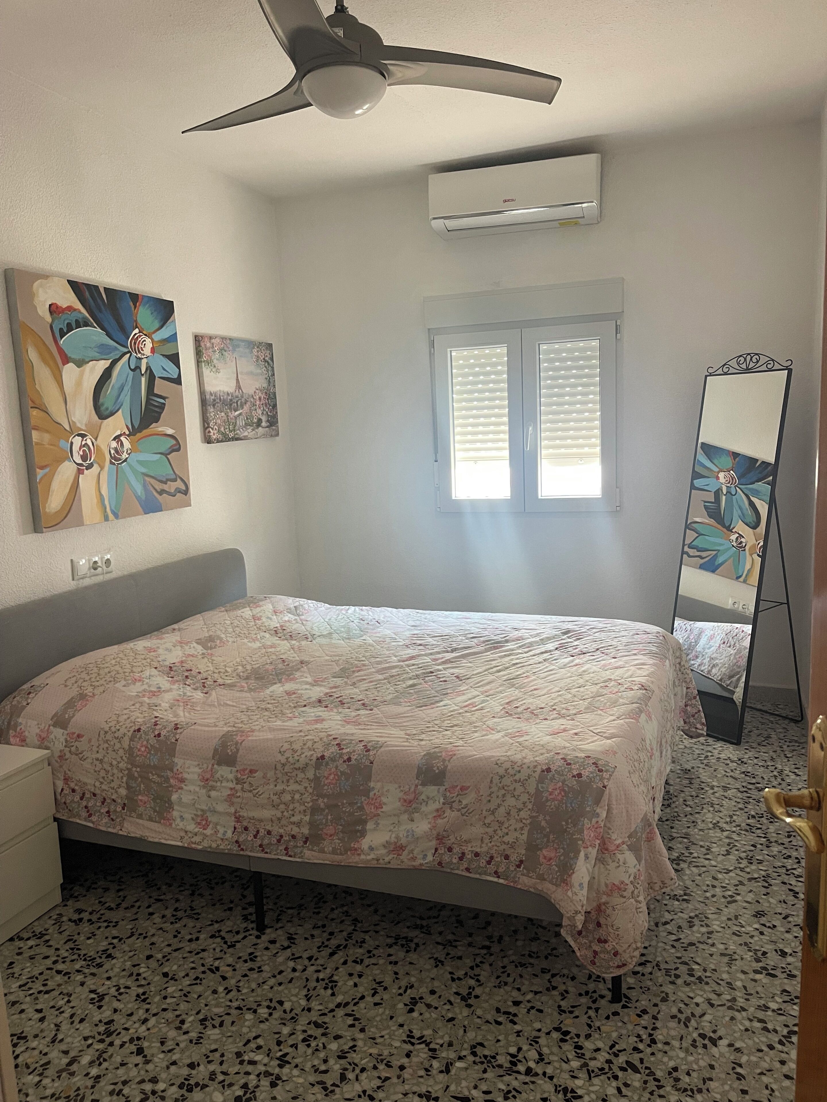 Une chambre avec un lit, un ventilateur de plafond, un appareil de climatisation, une fenêtre avec des stores et un miroir posé sur un support.