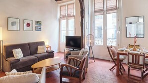 Een woonkamer met een bank, televisie, eettafel, stoelen en grote ramen.