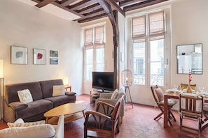 Ein Wohnzimmer mit Sofa, Fernseher, Esstisch, Stühlen und großen Fenstern.