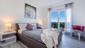A modern bedroom with a bed, bedside table, and a view of greenery through large windows. - Stunning home in Lumbarda (Lumbarda)