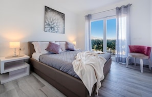 A modern bedroom with a bed, bedside table, and a view of greenery through large windows. - Stunning home in Lumbarda (Lumbarda)