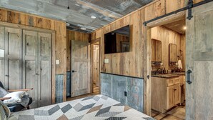 Ein rustikales Zimmer mit Holzwänden, einem Bett mit geblümtem Quilt, einem Stuhl und einem durchgehenden Türrahmen einsehbaren Badezimmer.