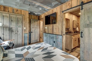 Ein rustikales Zimmer mit Holzwänden, einem Bett mit geblümtem Quilt, einem Stuhl und einem durchgehenden Türrahmen einsehbaren Badezimmer.
