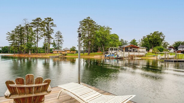 A wooden dock with a lounge chair and a wooden Adirondack chair, a small house with a dock, and a calm lake with trees. - Tiny Cabin on Lake Murray, Private Dock for Boats (Chapin)