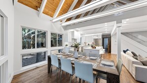 Eine moderne Küche mit weißen Schränken, einem Essbereich mit Holztisch und Blick ins Freie durch große Fenster.