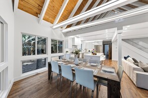Eine moderne Küche mit weißen Schränken, einem Essbereich mit Holztisch und Blick ins Freie durch große Fenster.