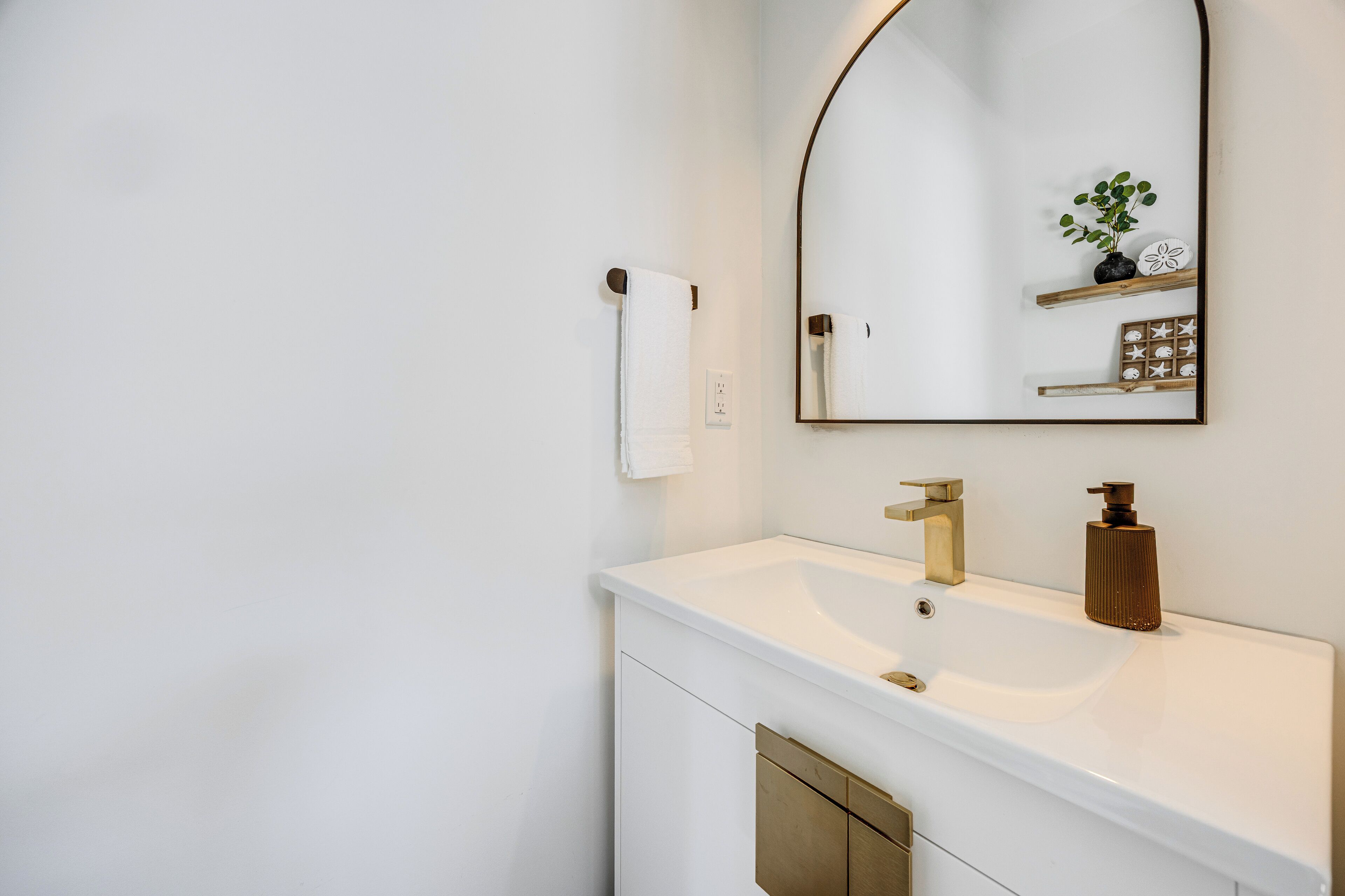 A modern bathroom with a white vanity, a gold faucet, a mirror with a wooden frame, and a small plant on the shelf.