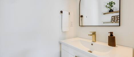 A modern bathroom with a white vanity, a gold faucet, a mirror with a wooden frame, and a small plant on the shelf.