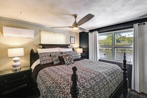 Un dormitorio con una cama grande, un ventilador de techo, un equipo de aire acondicionado, una ventana con vistas al agua y una mesita de noche con una lámpara.