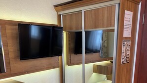 A compact room with a wooden cabinet, a television mounted on the wall, a small trash bin, and a chair. - KAAN AİLE PANSİYON (Amasra)