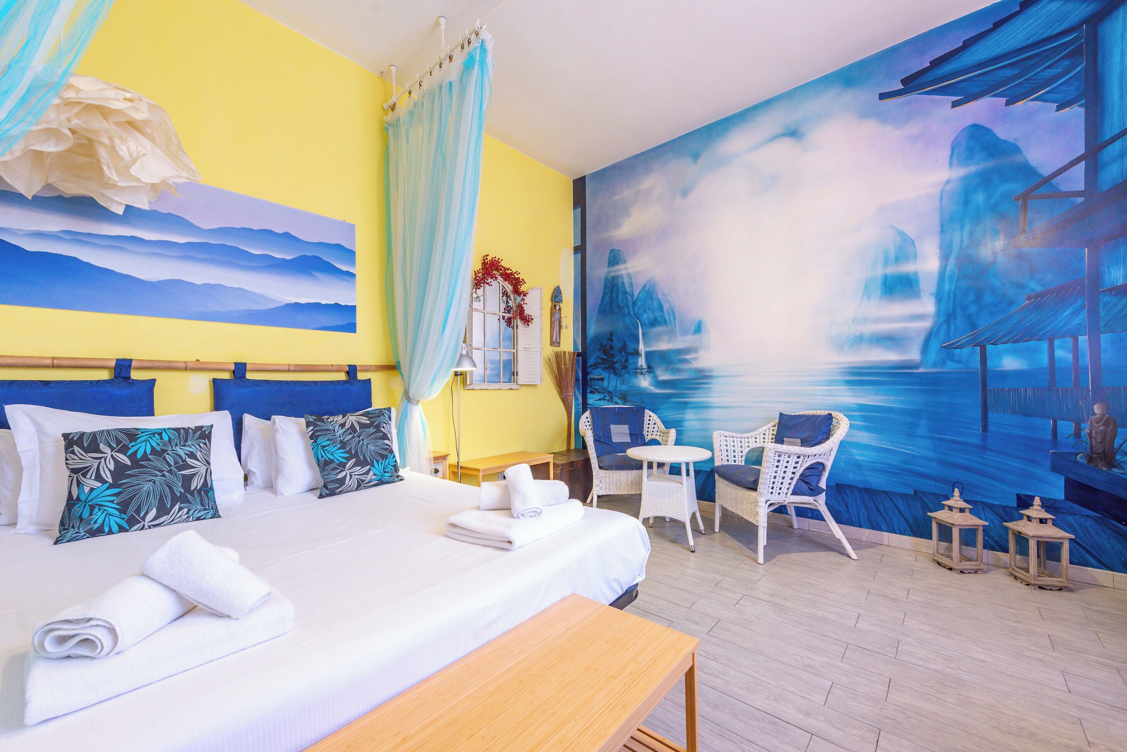 A hotel room with a bed, a chair, a small table, and a mural on the wall.