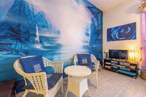 Une pièce avec un grand mur décoré d’une fresque, des meubles en osier, un téléviseur et une bibliothèque.