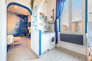 Ein kompakter Wohnraum mit Waschmaschine, einem kleinen Tisch und Blick auf die Stadt durch ein Fenster.