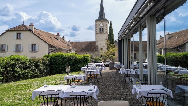 An outdoor dining area with tables and chairs, a clock tower in the background, and a building with large windows.