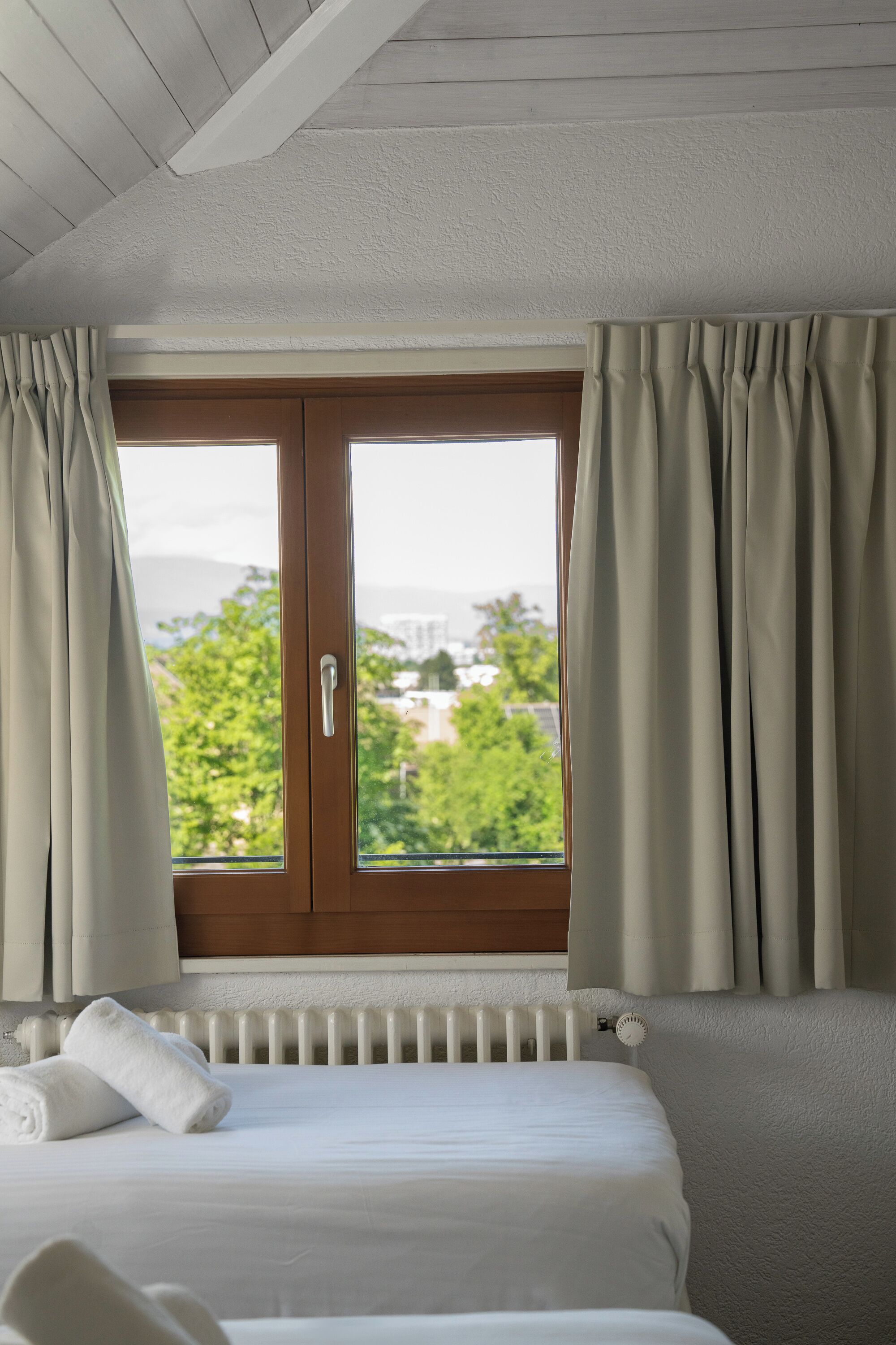 Ein Hotelzimmer mit Bett, einem Fenster mit Vorhängen und Blick auf Grünflächen und Gebäude.