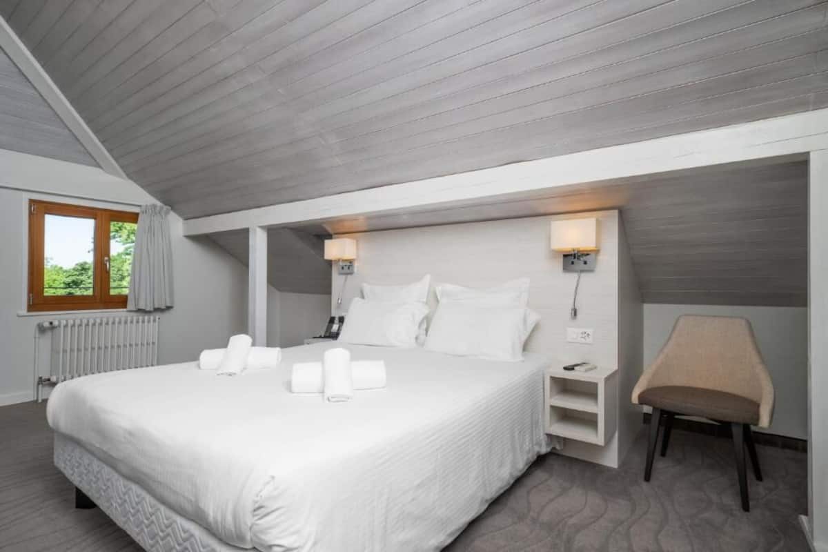 A modern hotel room with a large bed, bedside lamps, a chair, and a view of greenery outside.