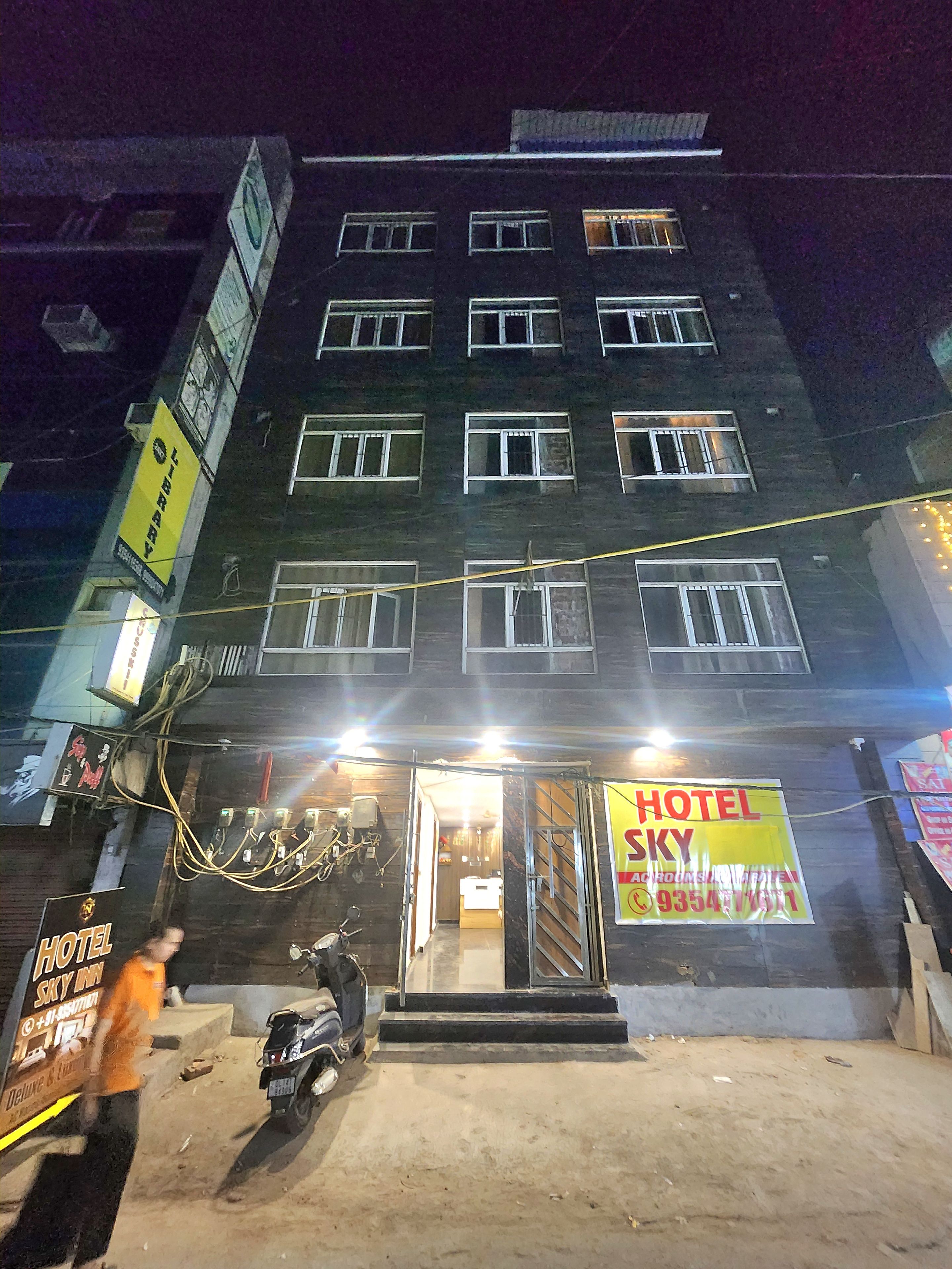 Un immeuble à plusieurs étages, sur lequel on peut voir une grande enseigne lumineuse indiquant « HÔTEL SKY », une moto garée devant, et une personne qui passe.