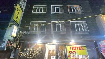 Un immeuble à plusieurs étages, sur lequel on peut voir une grande enseigne lumineuse indiquant « HÔTEL SKY », une moto garée devant, et une personne qui passe.
