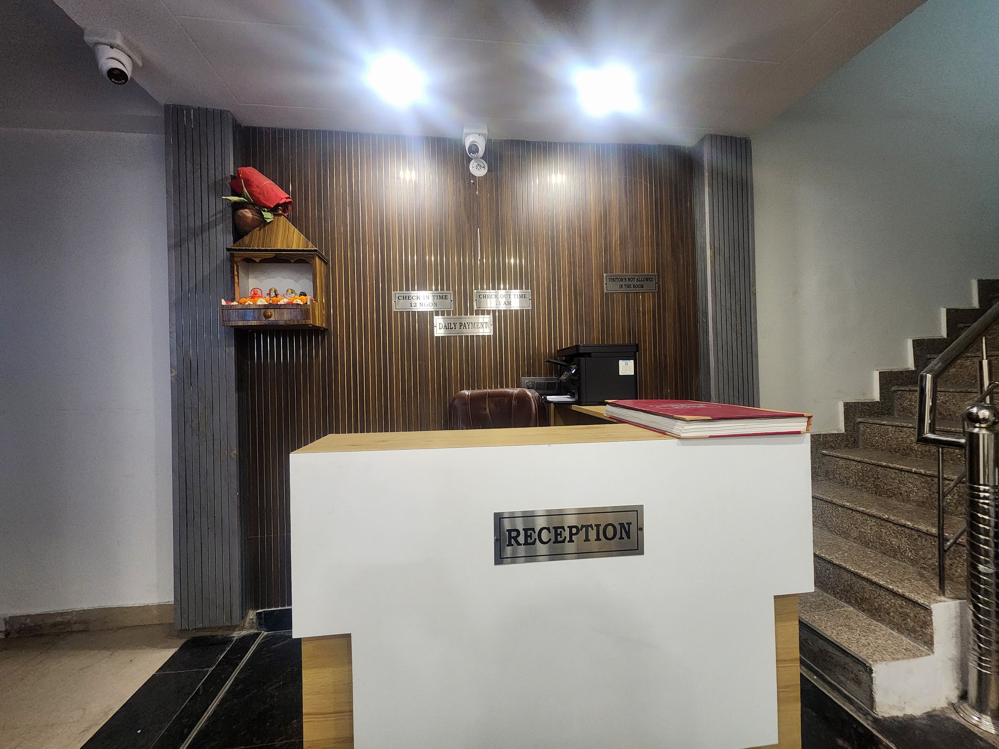 A reception desk with a 'RECEPTION' sign, a security camera, a 'CHECK IN TIME 12 NOON' sign, a 'DAILY PAYMENT' sign, and a staircase to the right.