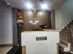 A reception desk with a 'RECEPTION' sign, a security camera, a 'CHECK IN TIME 12 NOON' sign, a 'DAILY PAYMENT' sign, and a staircase to the right. - Oly Hotel Sky Inn (New Delhi)