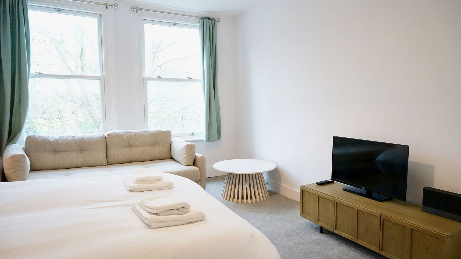 A hotel room with a large bed, a sofa, a small round table, a TV on a wooden cabinet, and a window with curtains.