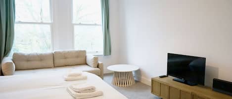 A hotel room with a large bed, a sofa, a small round table, a TV on a wooden cabinet, and a window with curtains.