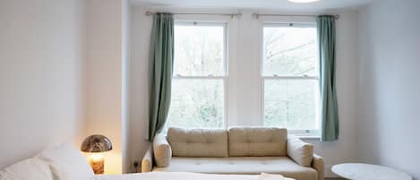 A bedroom with a bed, a sofa, a round table, and two windows with curtains.